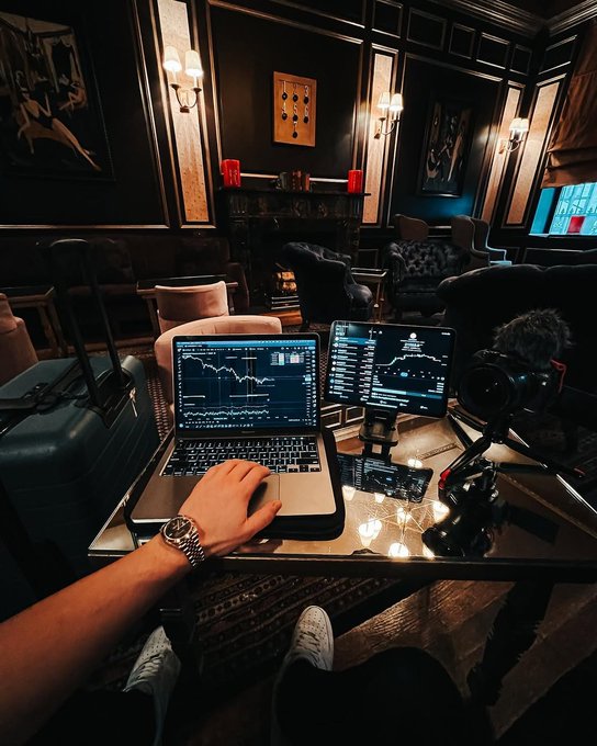 Person working on a laptop displaying trading charts and data, with an additional monitor showing analytics in a dimly lit, luxurious lounge setting.
