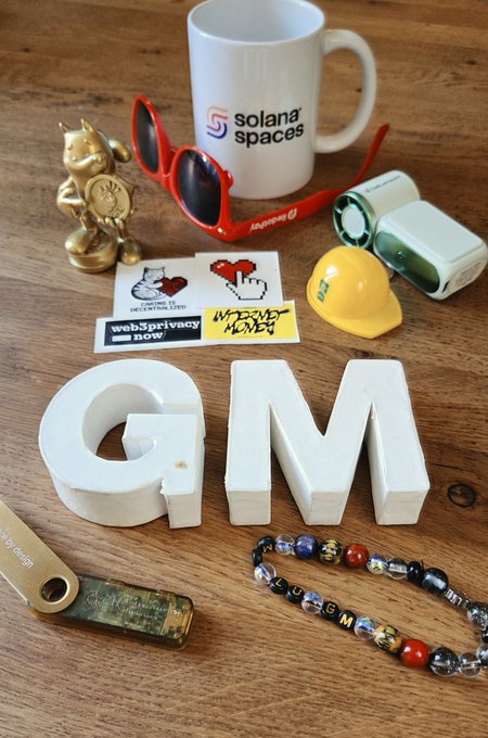 A 'Solana Spaces' branded mug surrounded by various items including red sunglasses, a gold cat figurine, stickers reading 'web3 privacy now', a 'GM' bracelet, and large 'GM' letters.