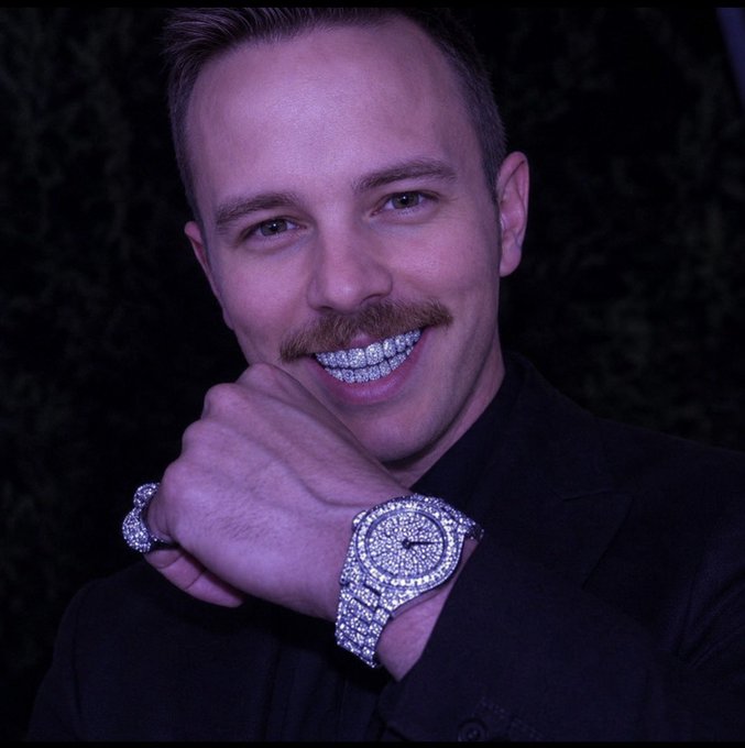 A smiling man with a mustache shows off a diamond-encrusted grill, ring, and luxury watch against a dark background.