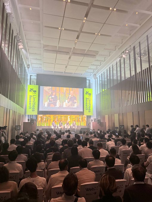 A large audience fills a modern conference hall with "Limitless Research" banners displayed on either side of a stage, where panelists are seated for a presentation.