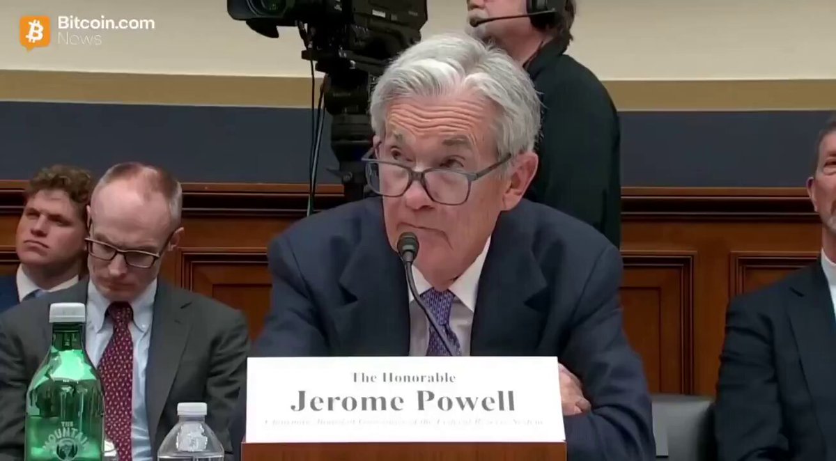 The image shows a man sitting at a desk, speaking into a microphone while wearing glasses. The background reveals that he is seated behind a podium with a sign displaying his name, "Powell". There are other people present in the room as well, suggesting this could be an event or gathering related to the former US President's activities. A bottle can also be seen on one of the tables nearby.