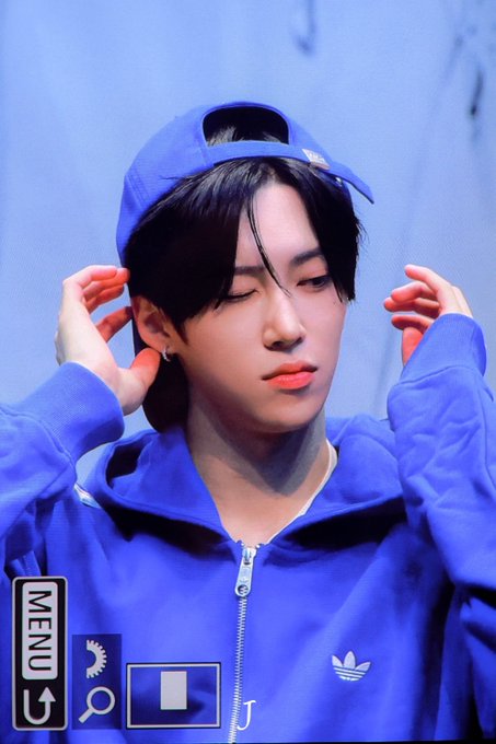 A young man with dark hair wears a blue Adidas track jacket and matching cap backwards, posing with his hands near his ears against a blue background. The image appears to be a photo taken at an event, with hashtags referencing the group ALPHADRIVEONE and member ARNO, suggesting this is fan-taken content from their "ALD1" promotion.
