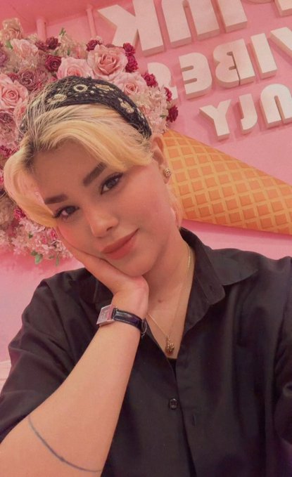 A stylish blonde woman with a black bandana poses in front of a pink wall featuring oversized white lettering and a floral display, while wearing a black shirt, watch, and delicate necklace. The vibrant pink setting and confident pose complement the motivational post's message of embracing a fresh week with bold energy and determination.