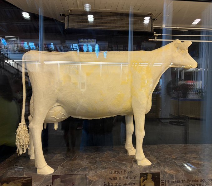 A life-sized sculpture of a yellow cow, made of butter according to the partially visible plaque crediting Garth Brooks, is displayed behind glass in what appears to be a hotel lobby.