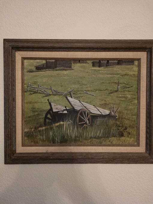 A framed landscape painting depicting an old wooden wheelbarrow sitting in a grassy field, with a rustic wooden fence and small buildings in the background.