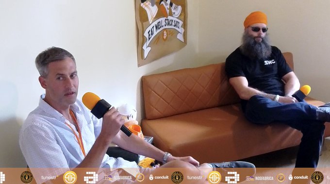 Two men sit on a brown leather couch during an interview at 'Eat Well, Stay Sill' bar, with one holding an orange microphone and logos for 'tunestr', 'ATL', 'conduit', and 'NOSFABRICA' displayed at the bottom.