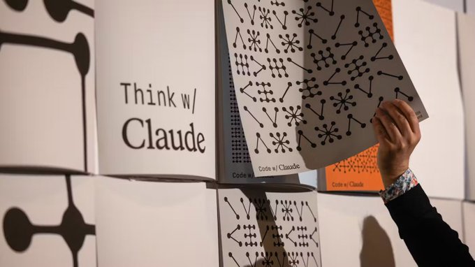 A person's hand lifts one of a stack of white "Think w/ Claude" and "Code w/ Claude" zine-style booklets, revealing decorative constellation-like patterns on the pages. The image illustrates Anthropic's Claude brand identity in the context of the company's massive new computing contract valued at up to 80 trillion yen, highlighting the scale of infrastructure investment behind its AI products.