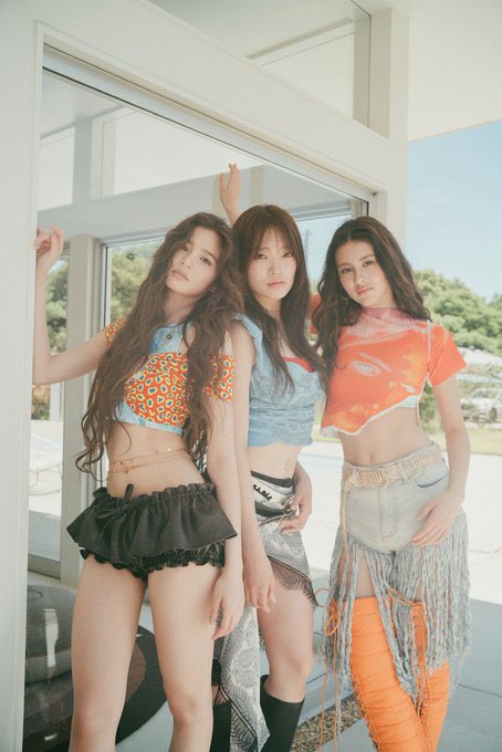 Three young women pose together in a sunlit doorway, wearing colorful crop tops and stylish skirts in orange, blue, and denim tones.