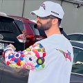 A low-resolution photo of a person wearing a white cap and t-shirt, holding what appears to be a colorful item, standing near a vehicle.