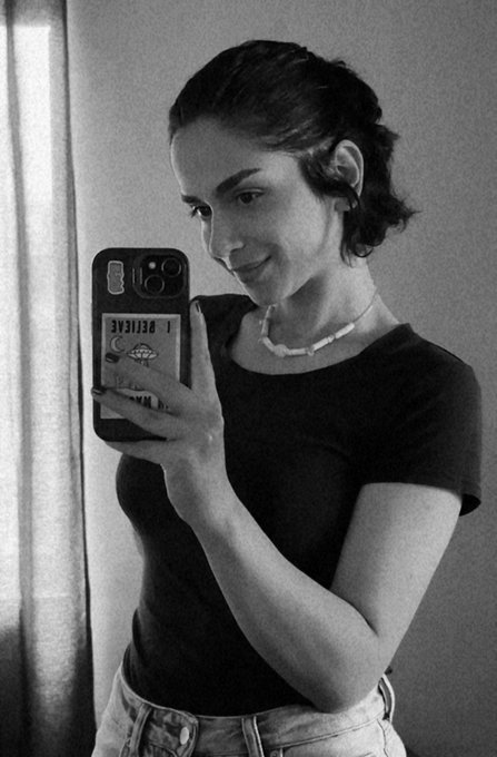 Black and white photo of a young woman taking a mirror selfie with a phone in a case that says 'I BELIEVE'.