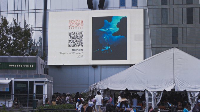 An outdoor scene showing a large billboard for 'BOOTZ CULT' featuring Ian Maina's artwork 'Depths of Wonder' (2022) with a QR code, displayed above a white event tent and a 'BURGERS+FRIES' food stand with people gathered below.