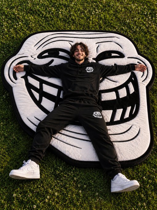 A young man lies smiling on a large black and white rug shaped like a grinning face mask, set on green grass, wearing a black hoodie and sweatpants with white sneakers.