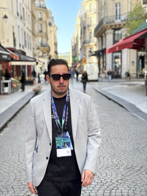 A man in a light gray blazer and sunglasses stands on a cobblestone Paris street wearing a Paris Blockchain Week lanyard with badge number 209.