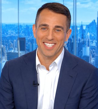 A smiling man in a navy blue suit and white shirt appears on a news set with a city skyline backdrop, with a small microphone clipped to his lapel. The image accompanies Anthony Pompliano's commentary criticizing U.S. monetary policy and predicting long-term Bitcoin price increases.