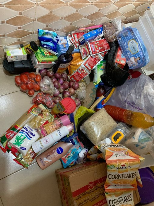 A haul of groceries including Nescafé coffee, Astra margarine, Nivea lotion, Dettol, Nasco Cornflakes, tomatoes, onions, garlic, and rice is piled on a tiled floor. The image accompanies a social media post celebrating that $50 earned online covered the cost of the shopping haul.