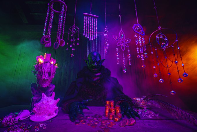 A goblin-like character wearing a green mask and dark fur cloak sits at a table stacked with gold coins, surrounded by colorful hanging crystals under neon purple and green lighting.