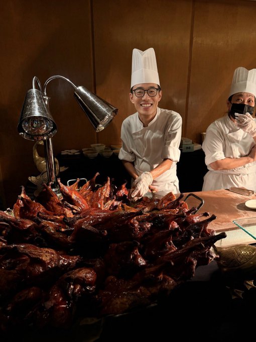 Two chefs in white uniforms and tall hats stand behind a large buffet display of roasted meats, with a heat lamp overhead and one chef smiling at the camera.