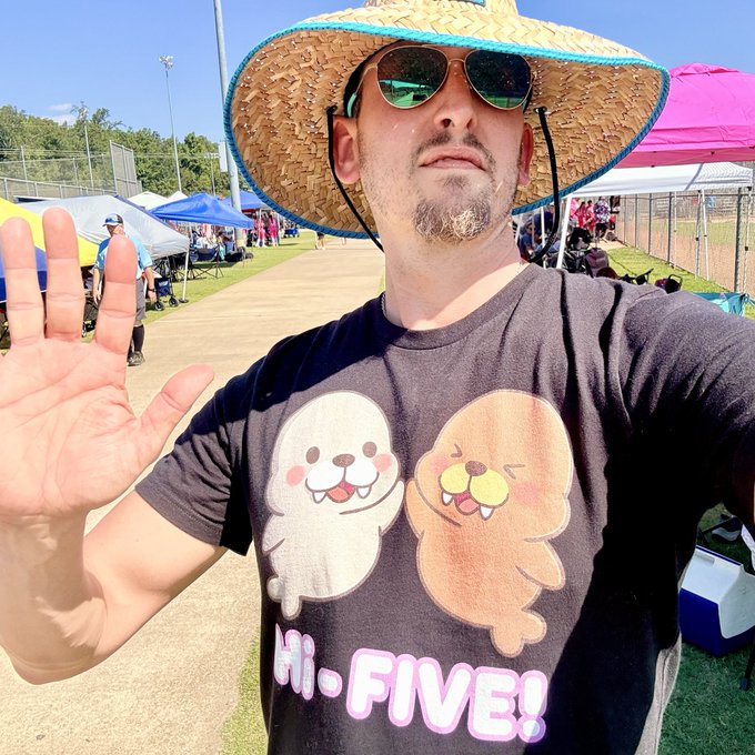 A man wearing a straw hat, sunglasses, and a t-shirt with cartoon sloths waves at the camera at an outdoor sports field. His shirt reads "Hi-FIVE!".