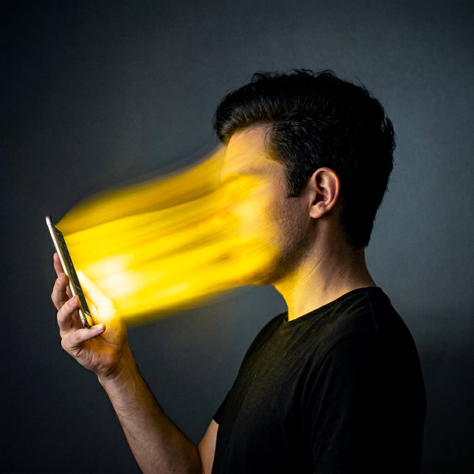 A man holds a phone in profile against a dark background while a glowing yellow-orange energy beam shoots from the screen into his face, visually embodying the caption's idea of "my phone and I merging into a single organism."