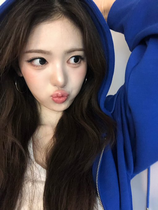 A young woman with long dark hair, wearing a blue hooded sweatshirt and silver hoop earrings, makes a pouty expression with her eyes looking to the side.