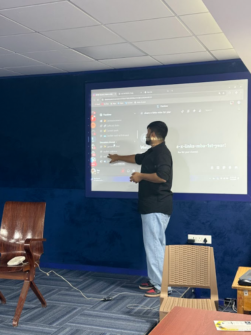 A young man in a black shirt and gray pants stands in front of a large interactive screen, pointing at a Google Drive interface showing a folder named 'e-x-links-mba-1st-year'.