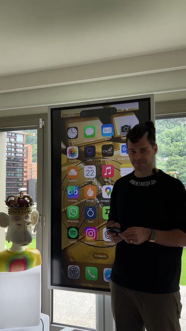 A man wearing a black t-shirt stands next to a large screen displaying an iPhone home screen with various app icons, while holding a phone in his hand.