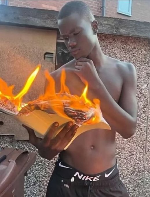 A shirtless young man wearing Nike shorts stands outdoors holding a book engulfed in flames, appearing to read it while fire spreads across the pages.