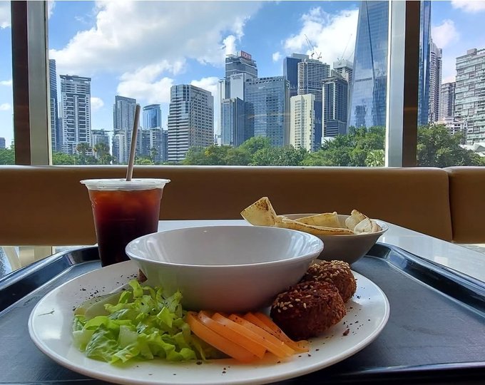 A meal of falafel, salad, pita bread, and iced tea served on a tray with a white bowl, set against a window view of a modern city skyline.