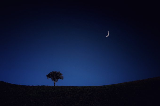 A serene night photograph showing a crescent moon hanging in a deep blue gradient sky above the dark silhouette of a lone tree on a hillside.