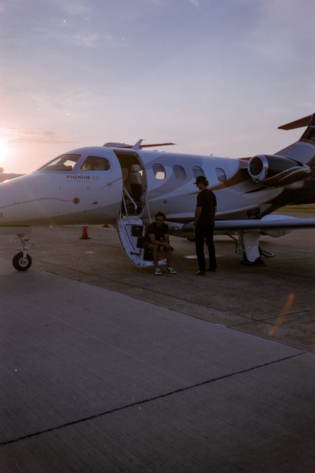 A man sits on the stairs of a private "PHENOM 100" jet at sunset, with another man standing beside him on the tarmac, suggesting the luxurious lifestyle following a "rainbet" sponsorship.