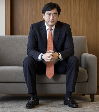 A professional photograph of an East Asian man wearing a dark suit and orange tie, sitting on a gray couch with his hands clasped together in a formal setting.