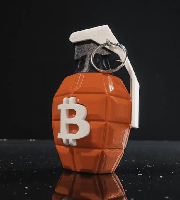 A 3D-rendered novelty keychain designed as an orange hand grenade with a white Bitcoin 'B' logo on its side sits against a dark, starry background.