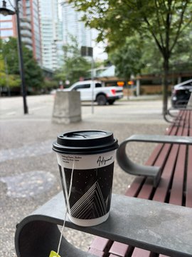 A black and white patterned coffee cup with a tea bag string hangs off a bench in an urban setting with buildings and a white pickup truck in the background.
