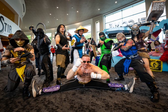 A group of cosplayers dressed as Mortal Kombat characters posing together at a convention, featuring characters like Scorpion, Sub-Zero, Raiden, and Shang Tsung in dynamic fighting stances.