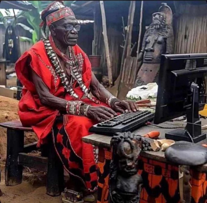 An elderly man wearing red traditional African attire and beaded necklaces sits at a wooden desk using a computer keyboard and monitor, with carved wooden sculptures displayed nearby.