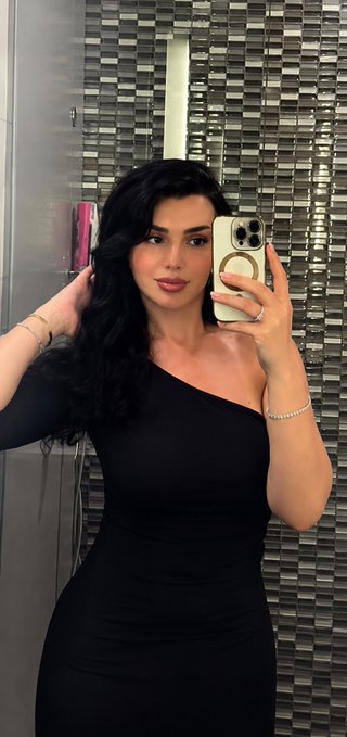 A woman with long dark hair takes a mirror selfie wearing a black one-shoulder dress, accessorized with a ring and bracelet.