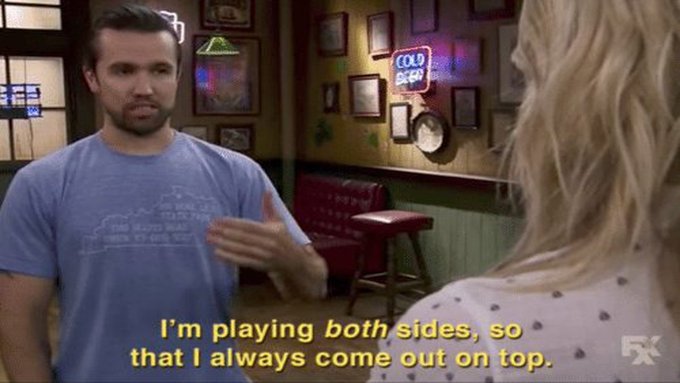 The image shows a scene from the FX TV show "It's Always Sunny in Philadelphia," featuring a bearded man in a blue t-shirt gesturing while speaking to a blonde woman in a dimly lit bar with neon signs and framed pictures on the walls. The subtitle reads "I'm playing both sides, so that I always come out on top," which humorously parallels the post's promotion of "The Black Bull token" as a strategic investment play.