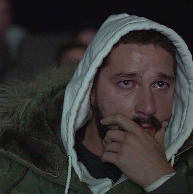 A close-up photograph of a man with a beard wearing a hooded jacket, resting his hand thoughtfully on his face with a serious, contemplative expression.