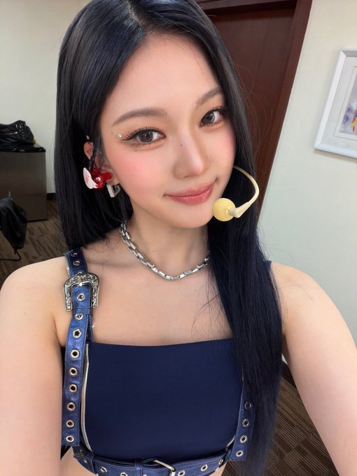 A young woman with long dark hair poses for a selfie wearing a dark blue top, denim overall straps with a rhinestone buckle, a silver chain necklace, and a headset microphone.