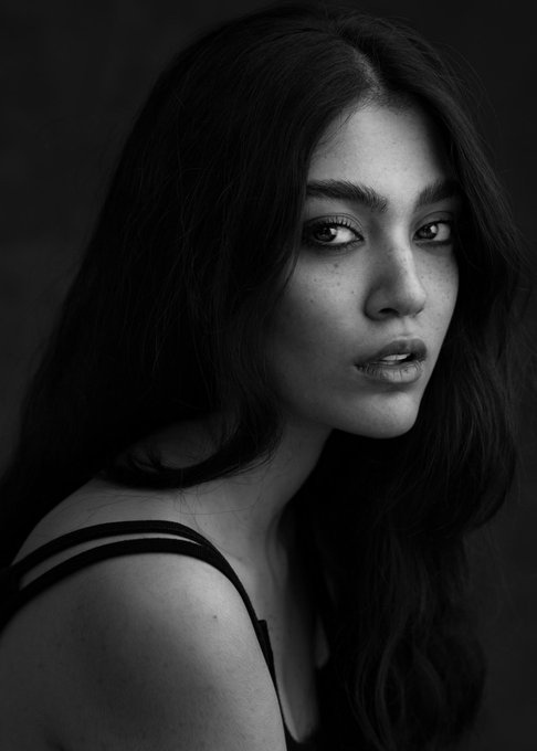 A black-and-white portrait photograph of a young woman with long dark hair looking over her shoulder, wearing a thin-strapped top against a dark background.