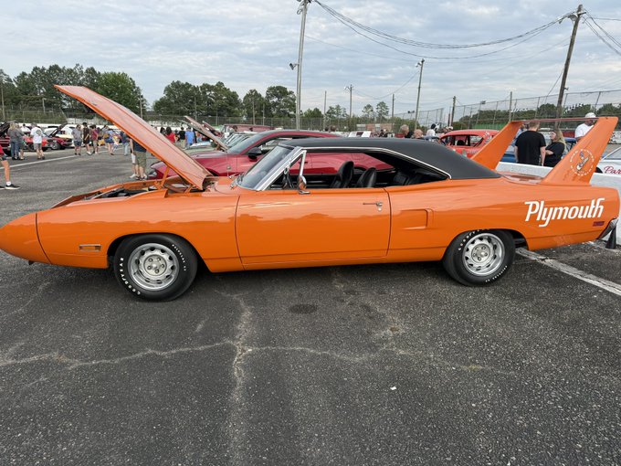 A bright orange 1970 Plymouth Superbird with its signature tall rear wing and "Plymouth" lettering displayed prominently is parked at what appears to be a car show. The image illustrates the post's reference to the $1 Doginal Dog token being valued at one of these iconic muscle cars.