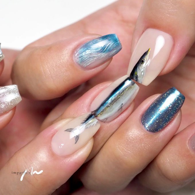 A close-up photograph of nail art featuring a fish design painted on long almond-shaped nails, with blue marbled and metallic blue accent nails, watermarked 'impartai'.