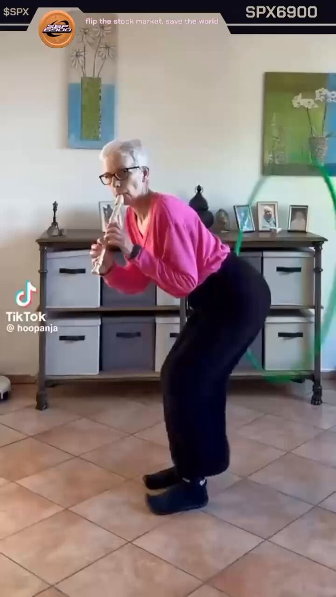 The image features an older woman wearing a pink shirt, black pants, and glasses standing on her tiptoes with one foot raised while holding a hula hoop. The background includes a white wall adorned with pictures and a wooden cabinet filled with various items such as vases and other decorations.