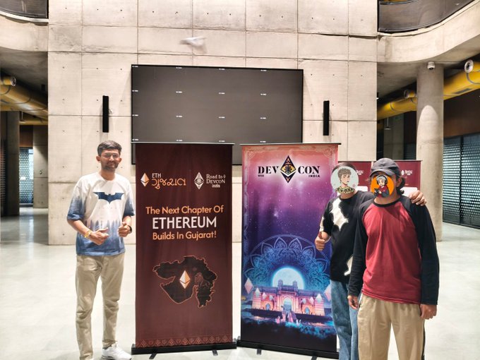 Three people pose in front of two promotional banners reading 'ETH 3.0/2.0' and 'DEVCON INDIA 2024' with the text 'The Next Chapter Of ETHEREUM Builds In Gujarat!', set in a modern concrete building interior.