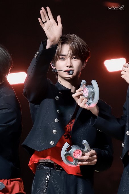 The image features a young man standing on stage, holding two game controllers with both hands raised high above his head. The background of the photo shows that he is part of a band performing at an event.