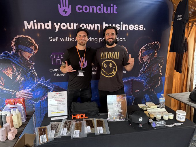 Two men stand behind a Conduit merchandise booth at what appears to be a convention, with promotional text 'Mind your own business. Sell without asking permission,' and products including 'Hot Flashes' jerky displayed on the table.