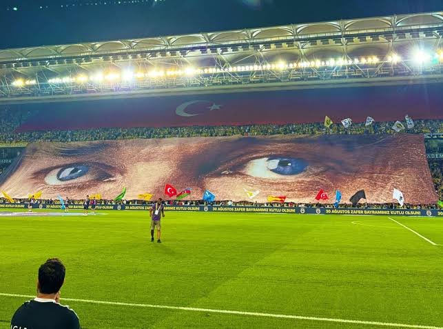 A massive tifo display featuring a pair of intense eyes with the Turkish flag and crescent moon unfurled in the stands, while a player stands alone on the pitch holding a Turkish flag. The banners lining the field read "BU AZUSTÜ ZAFER BAYRAMINA MUTLU OLUNUM" (Happy Victory Day Above This), connecting the "We Are Clean" caption to a message of patriotic celebration.