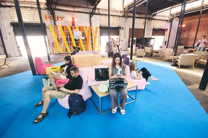 A group of people work and relax on colorful couches at the Devcon 8 conference in a large industrial-style venue with a "DEVCON" banner visible, illustrating the community atmosphere where developers discuss building open and verifiable computing systems rather than blindly trusting hardware and software. The casual setting reflects the collaborative exploration of how to make devices verifiable and trustworthy.