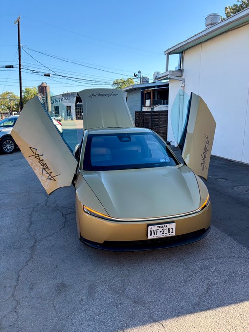 A gold/champagne-colored Tesla vehicle with its doors opened upward in a unique style, parked on a street with a Texas license plate reading 'XVF-3101'. The doors have stylized text/signature designs on them.