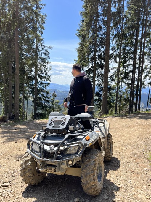 A man in a black jacket stands next to a muddy camouflage-patterned ATV in a forested mountain clearing with a scenic valley view behind him.